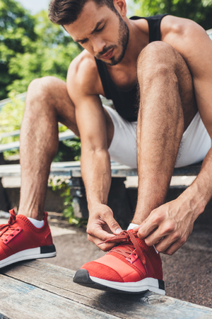 serious young sportsman tying shoelaces while sitting on bench at sport playgroundの写真素材