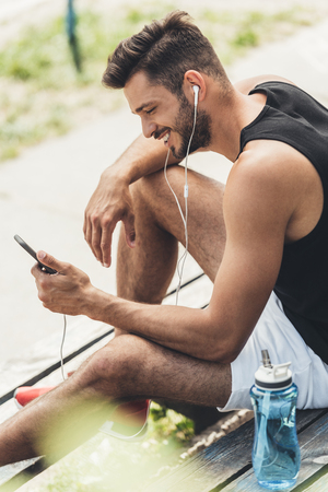 sportsman with bottle of water listening music with smartphone and earphones on bench at sport playgroundの写真素材