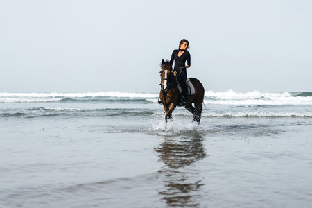 front view of young woman riding horse in wavy waterの写真素材
