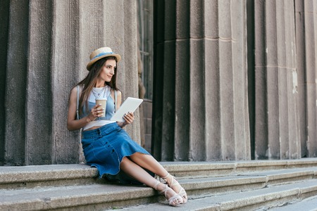 beautiful smiling young woman in hat holding paper cup and digital tablet while sitting on stairs and looking awayの写真素材