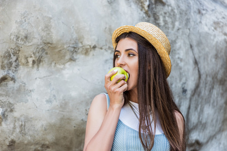 portrait of young stylish woman in hat eating fresh apple on streetの写真素材