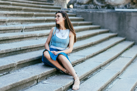 beautiful pensive young woman in eyeglasses sitting on stairs and looking awayの写真素材