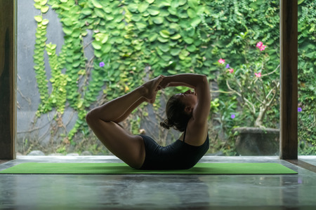 side view of sporty young woman practicing yoga in Bow pose (Dhanurasana) in front of wall covered...
