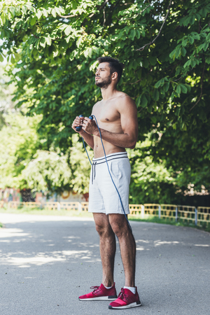 focused shirtless young man working out with jumping rope in front of green leavesの写真素材