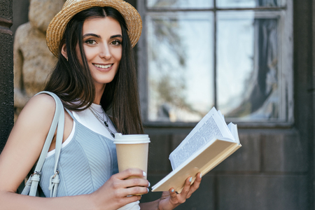 attractive girl with paper cup reading book and smiling at cameraの写真素材