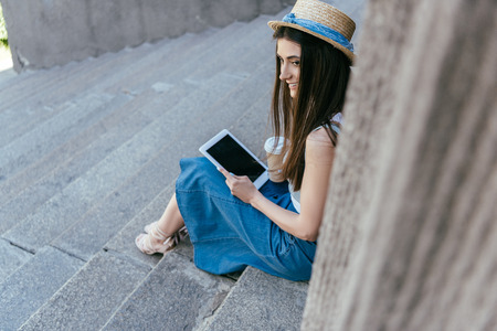 high angle view of smiling girl in hat holding paper cup and using digital tablet while sitting on stairsの写真素材