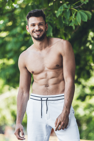happy shirtless young man looking at camera in front of green leavesの写真素材
