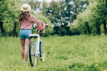 back view of young woman with retro bicycle with wicker basket full of flowers in forestの写真素材
