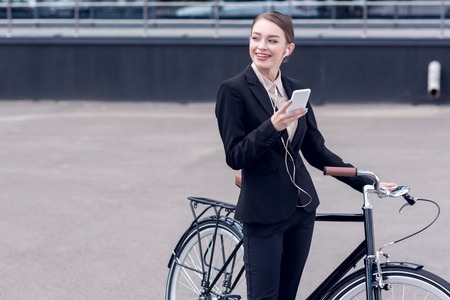 portrait of smiling businesswoman in earphones with smartphone standing near retro bicycle on streetの写真素材