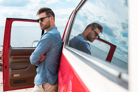handsome stylish male traveler in sunglasses posing at red car near the seaの写真素材
