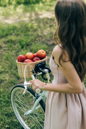 partial view of woman in dress holding retro bicycle with wicker basket full of ripe apples at countrysideの写真素材