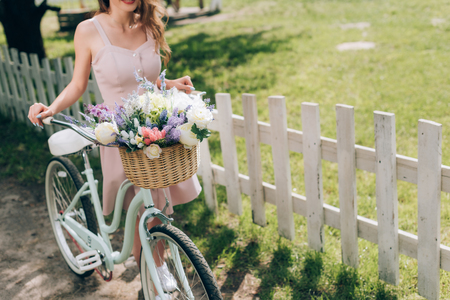 partial view of woman in stylish dress with retro bicycle with wicker basket full of flowers at countrysideの写真素材