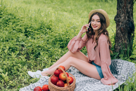 young smiling woman talking on smartphone while resting on blanket with wicker basket full of apples in parkの写真素材