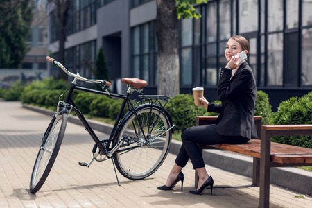young businesswoman with coffee to go talking on smartphone while resting on benchの写真素材