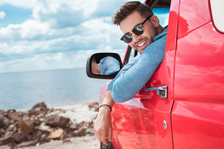 handsome cheerful man in sunglasses sitting in red car during road trip near the seaの写真素材