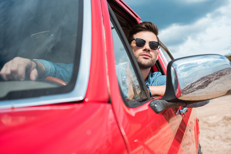 handsome man in sunglasses sitting in red car during road trip