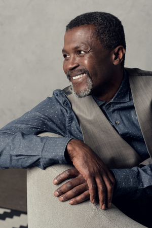 portrait of handsome smiling middle aged african american man in waistcoat sitting in armchairの写真素材
