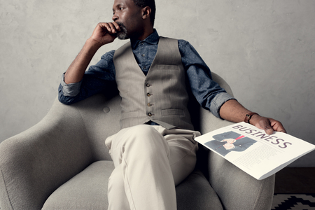 thoughtful african american man sitting in armchair with business newspaperの写真素材