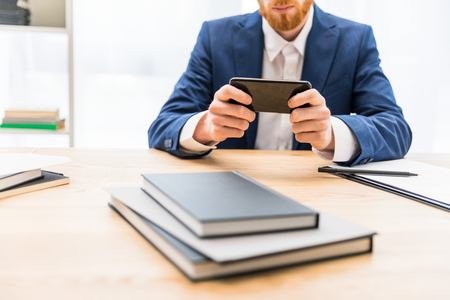 partial view of businessman in suit using smartphone at workplace with notebooks in officeの写真素材