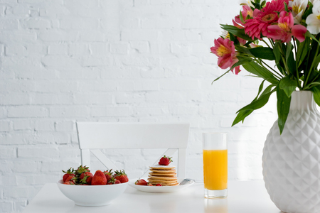 delicious pancakes with strawberries and orange juice on table with flowers in front of white brick wallの写真素材
