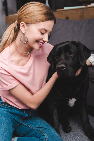 young smiling woman embracing black labrador on sofa at homeの写真素材