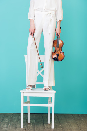 cropped view of girl holding violin and standing on chair, on blueの写真素材