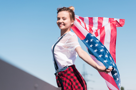 smiling young woman holding american flag against blue sky, independence day conceptの写真素材