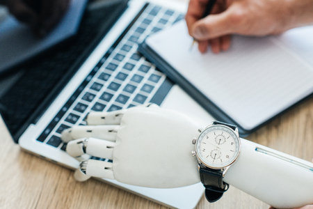 selective focus of robotic arm with wristwatch using laptop and human hand taking notes at wooden...