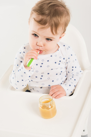 high angle view of baby boy eating puree from jar and sitting in highchair isolated on white backgroundの写真素材