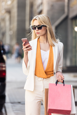 stylish blonde woman in sunglasses holding shopping bags and using smartphone on streetの写真素材