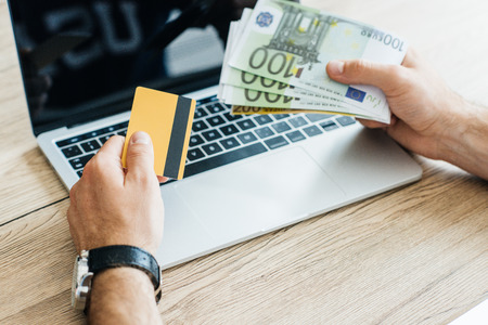 cropped shot of person holding credit card and euro banknotes above laptopの写真素材