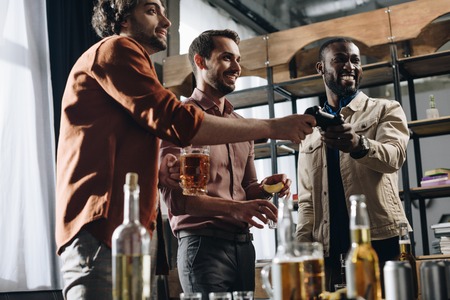 low angle view of smiling multiethnic men playing with joystick and drinking alcohol beverages togetherの写真素材