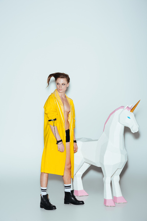 stylish man with hairstyle in yellow raincoat standing with big unicorn toy, on greyの写真素材