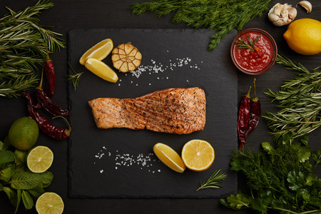 top view of grilled salmon steak with pieces of lemon and arranged ingredients around on black surfaceの写真素材