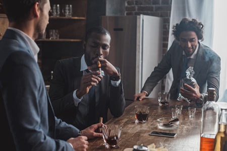 three multiethnic men in suits smoking cigars and drinking alcohol togetherの写真素材