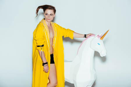 man with strange hairstyle in yellow raincoat standing with big unicorn toy, on greyの写真素材