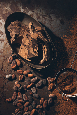 top view of delicious chocolate pieces, cocoa beans, powder and sieve on dark surfaceの写真素材