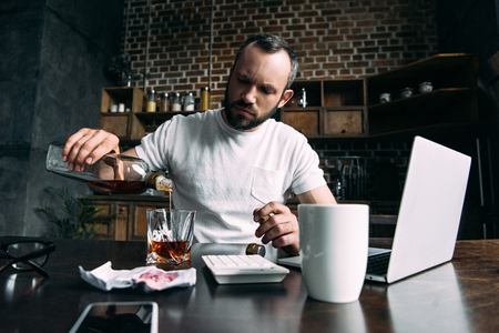 depressed young man pouring whiskey in glass after breakup with girlfriendの写真素材