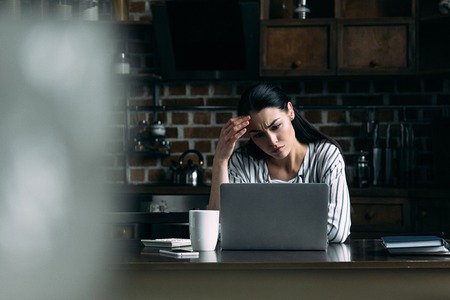 depressed young woman looking at laptop screen while sitting on kitchenの写真素材