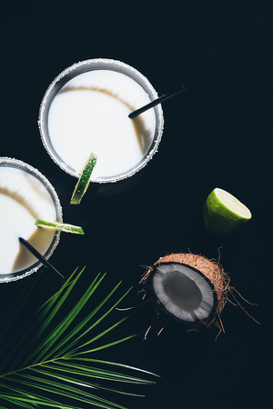 top view of glasses with coconut cocktails and drinking straws, lime and green palm leaves on blackの写真素材