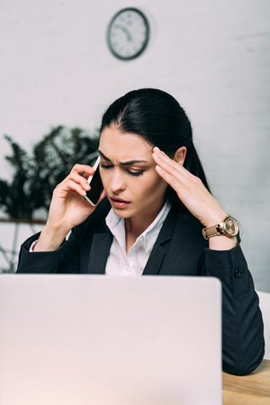 stressed businesswoman in suit talking on smartphone at workplace with laptop in officeの写真素材