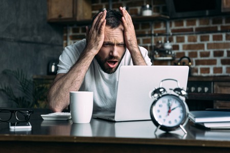 portrait of overworked freelancer remote working at table with laptop, clock and cup of coffee in kitchen at homeの写真素材