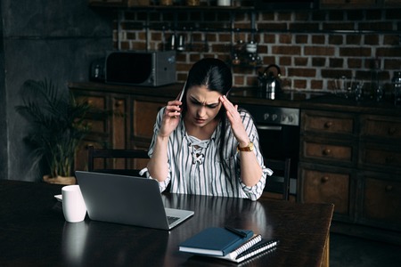 depressed young woman talking by phone and looking at laptop on kitchenの写真素材