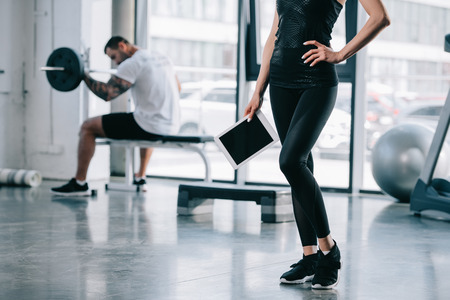 cropped shot of female personal trainer with digital tablet and athletic man exercising with barbell at gymの写真素材