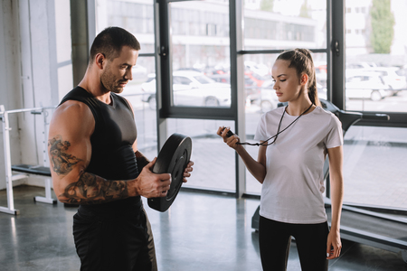female personal trainer using timer while sportsman doing exercises with barbell disc at gymの写真素材