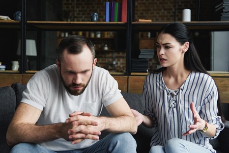 depressed young man sitting on couch and looking away while his wife pressing himの写真素材