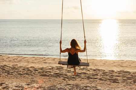 back view of woman on swing at ocean beach during sunsetの写真素材