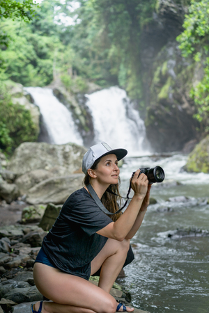 side view of woman in cap with photo camera in hands and Aling-Aling Waterfall on backdrop, Bali, Indonesiaの写真素材