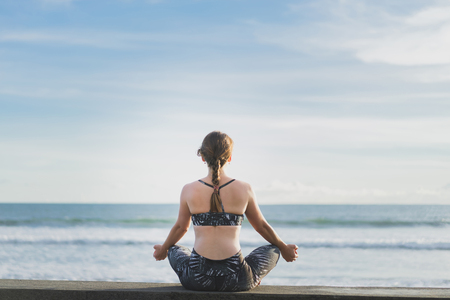 back view of woman meditating in lotus position with ocean and blue sky on background, Bali, Indonesiaの写真素材