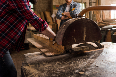 cropped shot of carpenter in checkered shirt using machine saw and partner standing behind at sawmillの写真素材
